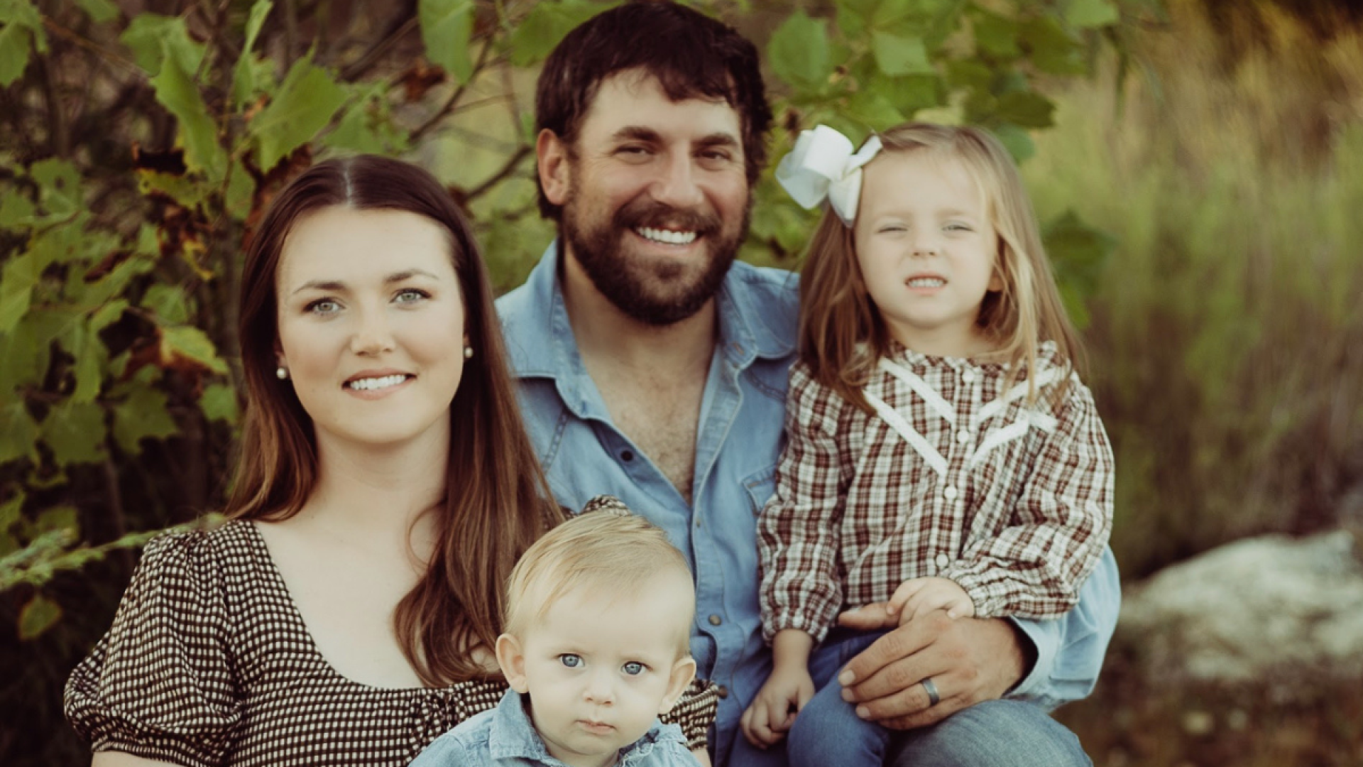 Husband and wife pose outdoors with their two young children, a toddler boy and a preschool-age girl with a white bow in her hair, surrounded by greenery.